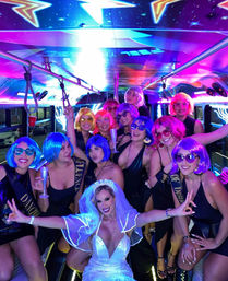 Bachelorette party on a neon-lit party bus — bride in a white gown surrounded by friends in colorful wigs, sunglasses and holding champagne flutes