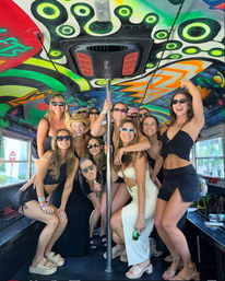 Group of friends in swimsuits and summer outfits posing around a chrome pole inside a colorful, psychedelic-painted party bus interior, wearing sunglasses and smiling for a lively daytime celebration.