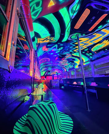 Neon blacklight party bus interior with fluorescent psychedelic ceiling mural, chrome dance pole, and striped glowing hats lined along the bench — vibrant UV-lit nightlife scene.
