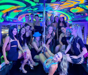 Group of women partying inside a neon-lit party bus with a chrome dance pole, colorful blacklight murals, drinks, and smiling faces, urban nightlife vibe.