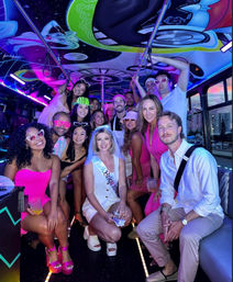 Group of friends on a neon-lit party bus for a bachelorette celebration, bride wearing sash and veil center-front, colorful mural ceiling, LED lights, playful sunglasses and drinks, everyone smiling for a group photo.