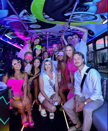 Group of friends on a neon-lit party bus for a bachelorette celebration, bride wearing sash and veil center-front, colorful mural ceiling, LED lights, playful sunglasses and drinks, everyone smiling for a group photo.