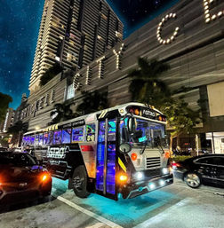 Neon-lit retro party bus labeled ASTRO with blue underglow and colorful interior lights parked on a downtown city street at night beside palm trees and a lit high-rise