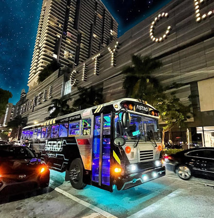 Neon-lit retro party bus labeled ASTRO with blue underglow and colorful interior lights parked on a downtown city street at night beside palm trees and a lit high-rise