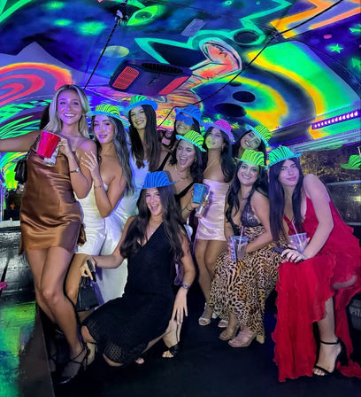 Group of women in cocktail dresses posing inside a neon-lit party bus with psychedelic ceiling art, wearing striped glow hats and holding drinks for a lively night-out celebration.