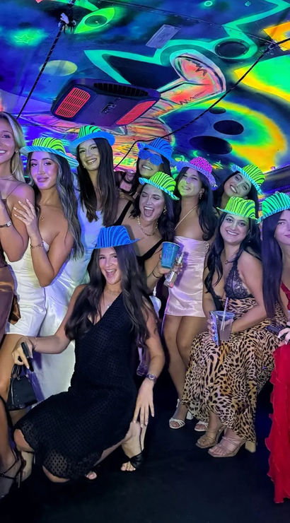 Group of women in cocktail dresses posing inside a neon-lit party bus with psychedelic ceiling art, wearing striped glow hats and holding drinks for a lively night-out celebration.