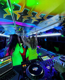 Neon-lit party bus interior at night with DJ turntables and laptop in the foreground, colorful ceiling graphics, and a crowd in bright outfits dancing and holding overhead rails.