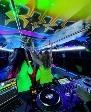 Neon-lit party bus interior at night with DJ turntables and laptop in the foreground, colorful ceiling graphics, and a crowd in bright outfits dancing and holding overhead rails.