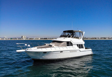 White motor yacht floating on deep-blue ocean near a sandy coastline and low-rise city skyline under a clear blue sky