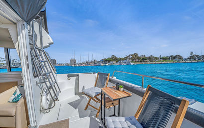 Sunny yacht aft deck with cushioned chairs and small wooden table overlooking turquoise marina and distant harbor skyline