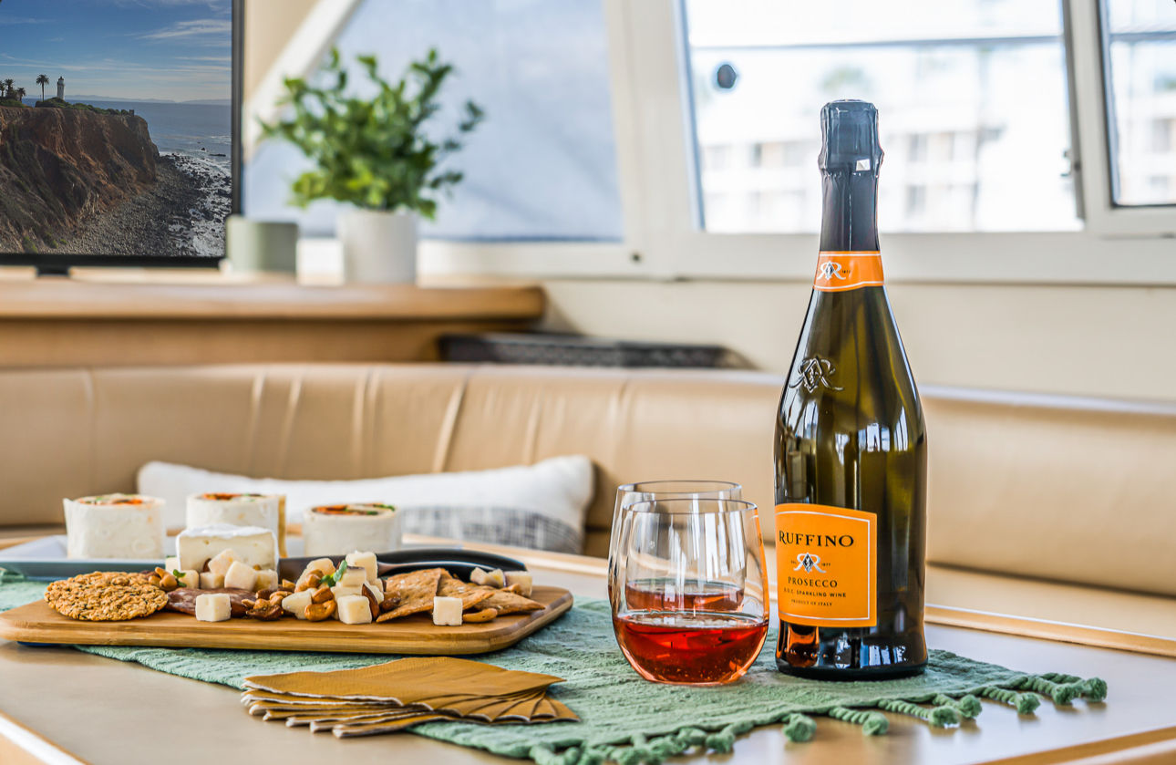 Sunlit yacht cabin scene with a Prosecco bottle and stemless glass of rosé beside a charcuterie board on a green runner, with a coastal cliff view through the window.