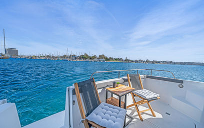 Two cushioned wooden chairs and a small side table on a white boat deck overlooking a bright blue harbor and marina with sailboats and shoreline under a clear sunny sky.