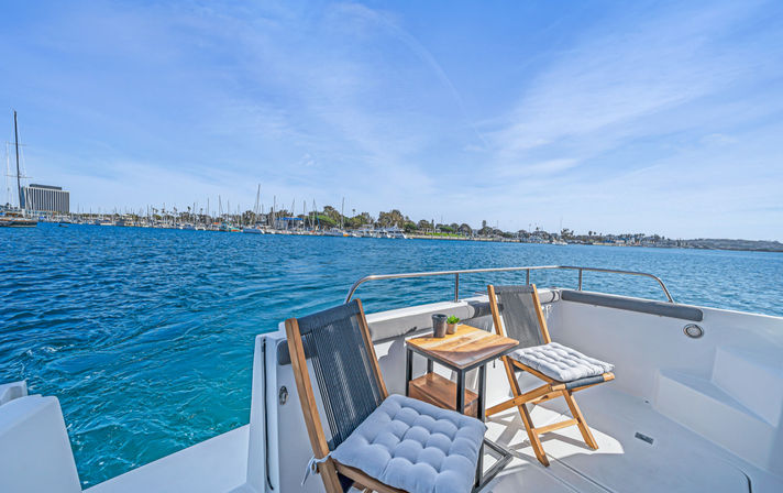 Two cushioned wooden chairs and a small side table on a white boat deck overlooking a bright blue harbor and marina with sailboats and shoreline under a clear sunny sky.