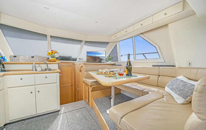 Bright luxury yacht interior with beige leather U-shaped dinette and wooden table set with wine and snacks, compact white galley cabinets, vase of sunflowers, and panoramic windows showing a blue ocean view.