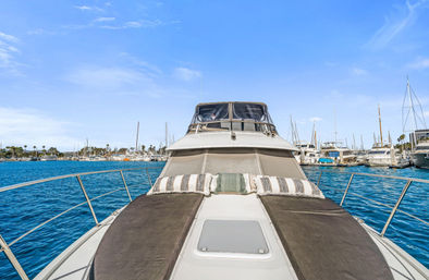 Bow of a luxury yacht with cushioned sun pads and striped pillows, surrounded by turquoise water and sailboats in a bustling coastal marina under a bright blue sky