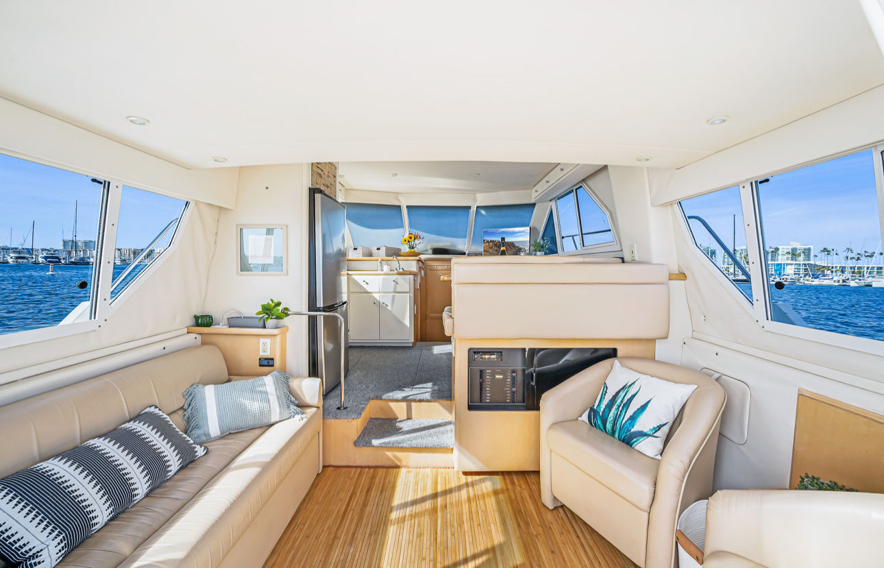 Sunlit yacht salon with cream leather seating, modern galley, bamboo floor and panoramic windows showcasing a blue marina with sailboats.