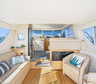 Sunlit yacht salon with cream leather seating, modern galley, bamboo floor and panoramic windows showcasing a blue marina with sailboats.