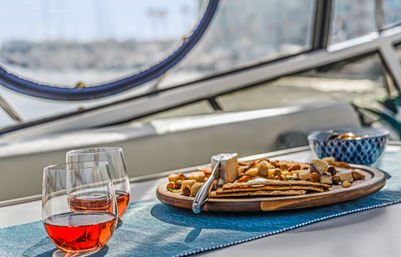 Two stemless glasses of rosé and a wooden charcuterie board with cheese, crackers, nuts and olives on a blue table runner inside a yacht, marina visible through a porthole.