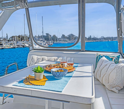 Sunlit yacht dining nook with white cushions and pillows, table set with bowls, snacks, wine and a small succulent, overlooking a blue marina filled with sailboats and a coastal harbor.