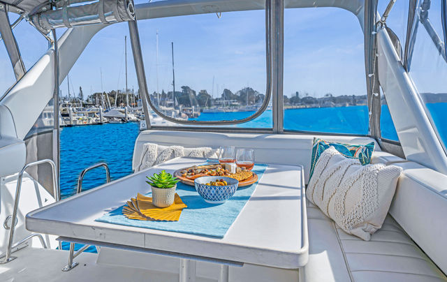 Sunlit yacht dining nook with white cushions and pillows, table set with bowls, snacks, wine and a small succulent, overlooking a blue marina filled with sailboats and a coastal harbor.