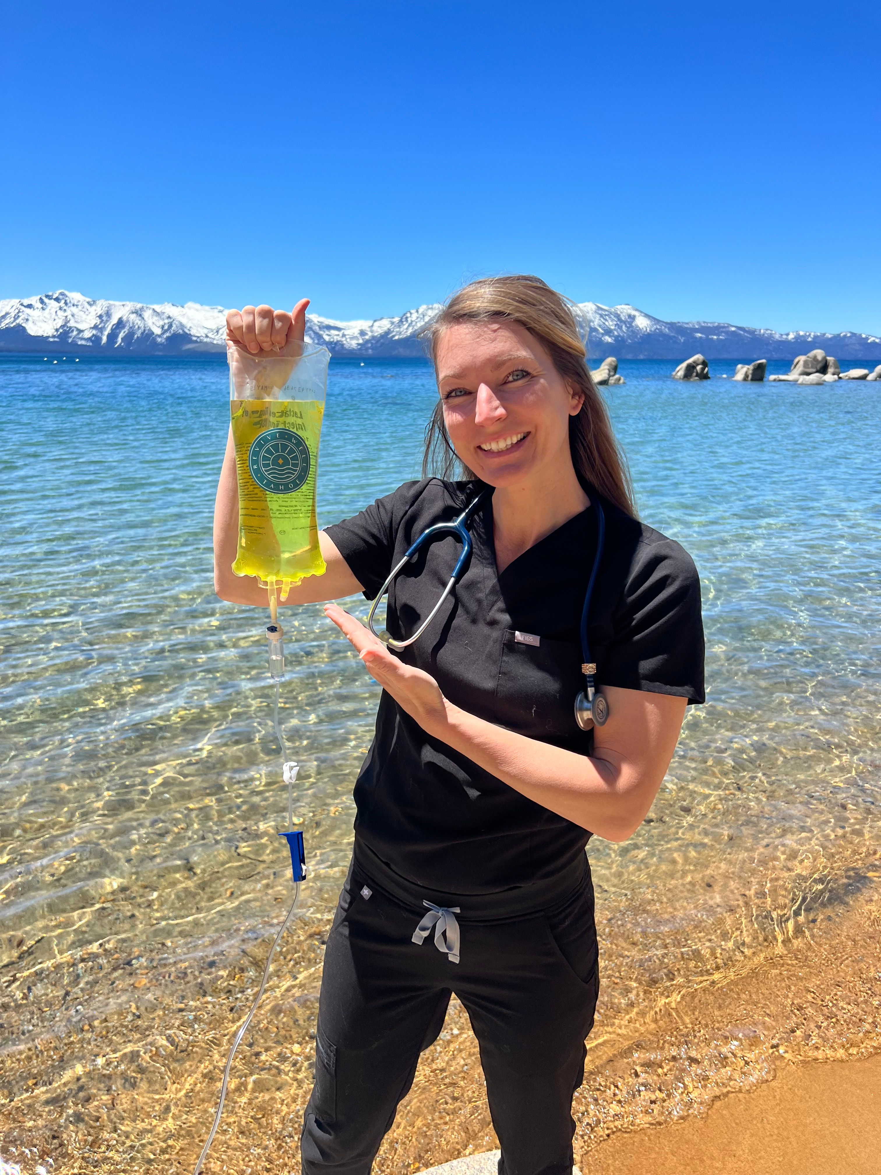 Medical professional in black scrubs with a stethoscope holding a yellow IV bag on a sunny alpine lake beach with clear water and snow-capped mountains