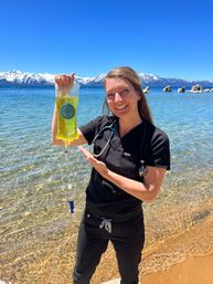Medical professional in black scrubs with a stethoscope holding a yellow IV bag on a sunny alpine lake beach with clear water and snow-capped mountains