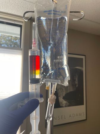 Gloved hand holding a syringe filled with red-to-yellow medication beside a clear IV saline bag hanging from a pole in a sunlit patient room, window and framed black-and-white landscape print in the background.