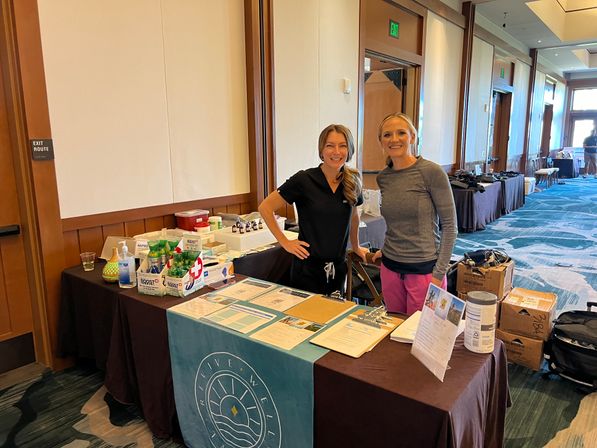 Two smiling women standing behind a wellness expo table with brochures, product samples and boxes set up in a hotel conference room.