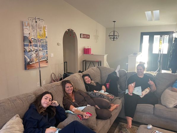 Four women lounging on a sectional couch in a bright living room receiving wellness IV drips from poles - cozy at-home IV drip party.