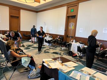 Plasma/blood donation drive in a hotel conference room with medical staff attending reclining donors, IV bags on stands, and clipboards and paperwork on nearby tables.