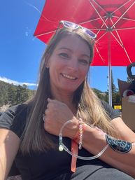 Smiling woman selfie outdoors under a red patio umbrella with sunglasses on her head, showing an IV hydration tube and beaded bracelets on her wrist, blue sky and pine-covered hillside in the background