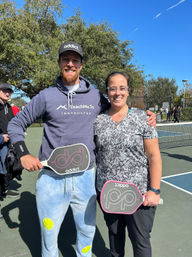 Two smiling pickleball players posing with paddles on a sunny outdoor court in a park, trees and blue sky behind them.