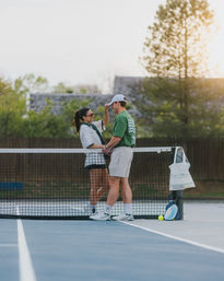 Two people on an outdoor tennis court at sunset, leaning on the net and smiling with a racket and tennis ball resting by the post.