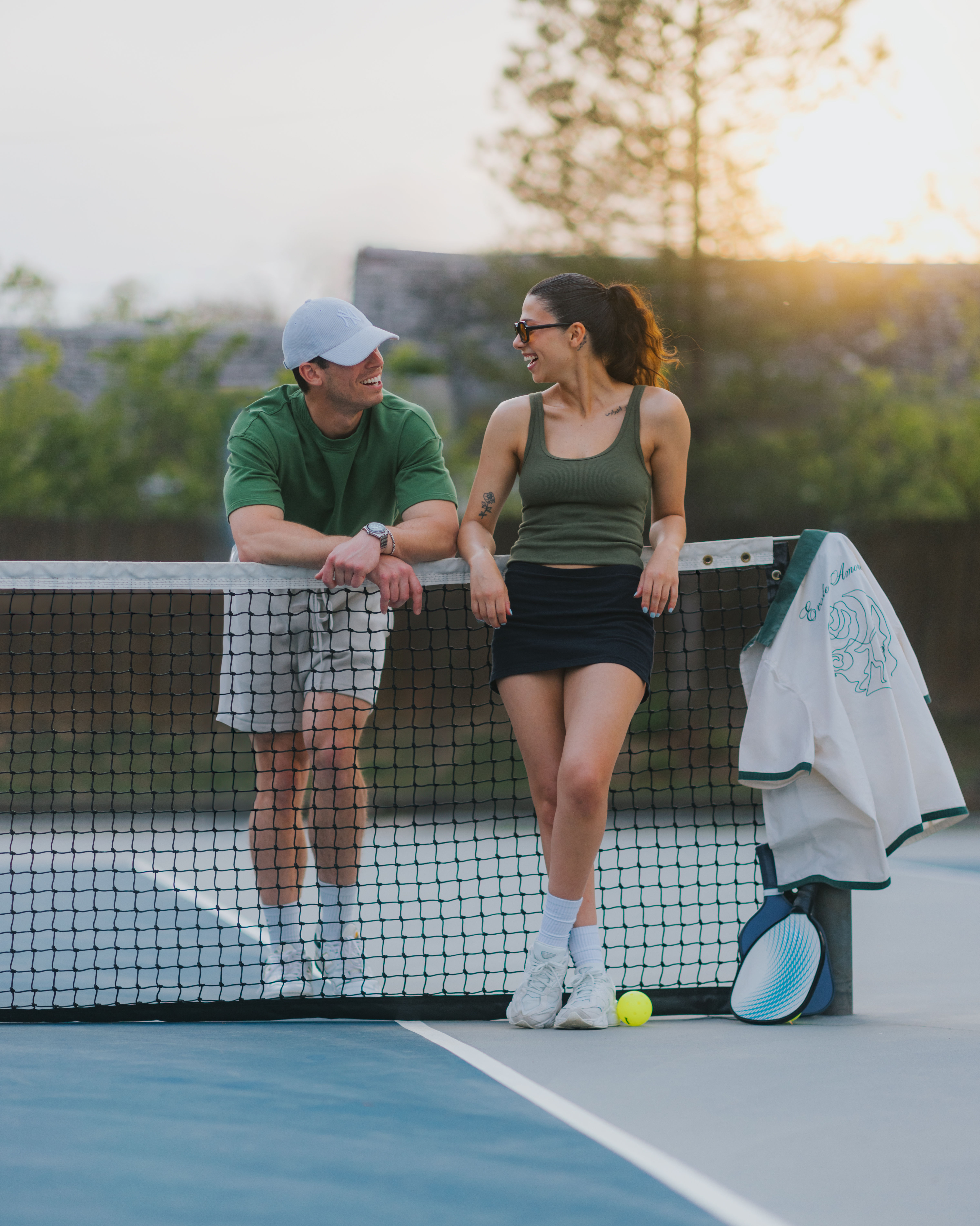 Two tennis players leaning on the net at an outdoor court at sunset, smiling and chatting with a racket, tennis ball, and jacket resting on the post.