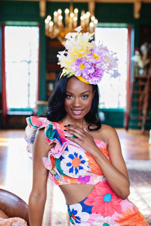 Smiling woman in a colorful one-shoulder floral dress wearing a bold purple-and-yellow flower headpiece, posing in an elegant room with a chandelier and tall windows — vibrant fashion portrait.