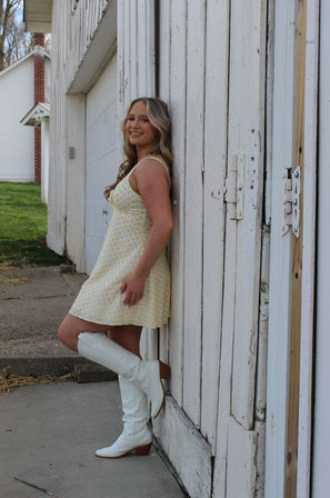 Outdoor portrait of a smiling woman in a yellow polka-dot dress and white knee-high boots leaning against a weathered white barn door at a farmhouse