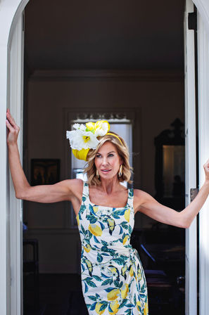 Sunlit doorway portrait of a woman in a lemon-print summer dress and oversized yellow floral headpiece, striking an elegant pose — spring/summer fashion.