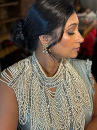 Close-up profile of a woman in a sleek low bun wearing a dramatic multi-strand pearl bib necklace, jeweled gold floral earrings and a nose stud — bridal glam updo.