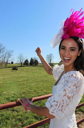 Smiling woman in a white lace dress and bright pink feather fascinator leaning on a red fence, raising her arm in a sunny rural pasture with grazing horses under a clear blue sky.