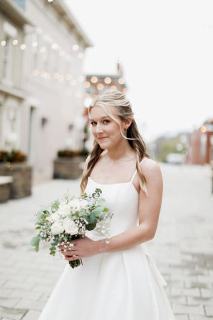 Smiling bride in a simple white gown holding a white rose and eucalyptus bouquet on a cobblestone downtown street with twinkling string lights