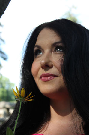 Close-up sunlit outdoor portrait of a woman with long dark hair holding a yellow wildflower near her chin and gazing upward with a dreamy expression.