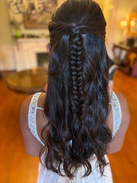 Back view of long dark hair in a half-up style with a central braid and loose waves, paired with a white beaded dress in a warm indoor living room