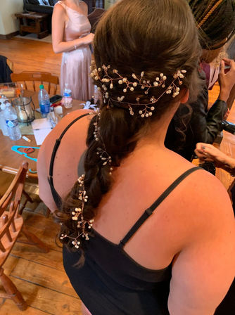 Bridal-style braided hairstyle woven with a delicate pearl-and-crystal vine, back view of a woman in a black camisole in a rustic room with wooden floors and a table of getting-ready items