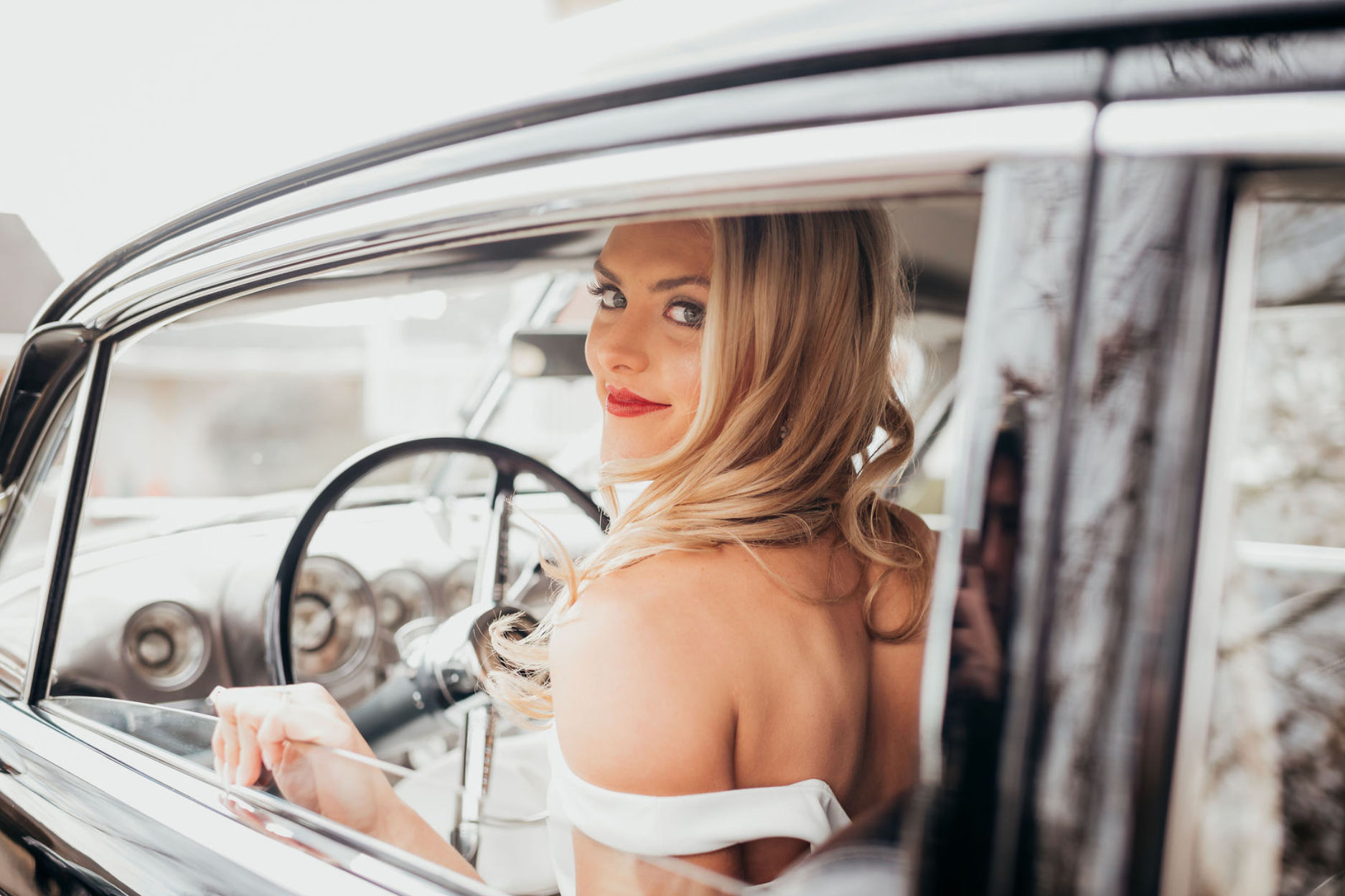 Blonde woman in an off-shoulder white dress smiling over her shoulder inside a shiny vintage car, showcasing the classic steering wheel and dashboard — retro glam portrait.