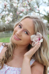 Smiling blonde woman in a pastel sundress holding a pink magnolia blossom to her ear in a spring orchard of blooming magnolia trees