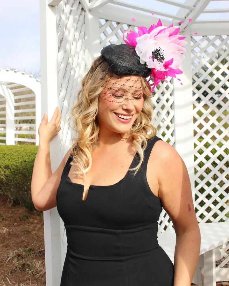 Smiling woman in a sleeveless black dress wearing a black fascinator with a pink-and-white flower and net veil, posing by a white lattice garden gazebo on a sunny day.
