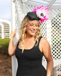 Smiling woman in a sleeveless black dress wearing a black fascinator with a pink-and-white flower and net veil, posing by a white lattice garden gazebo on a sunny day.