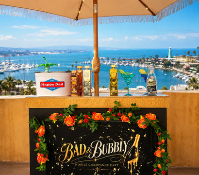 Rooftop mobile champagne cart decorated with orange roses and colorful bottles under a fringed umbrella, overlooking a sunny marina, harbor boats and a coastal lighthouse