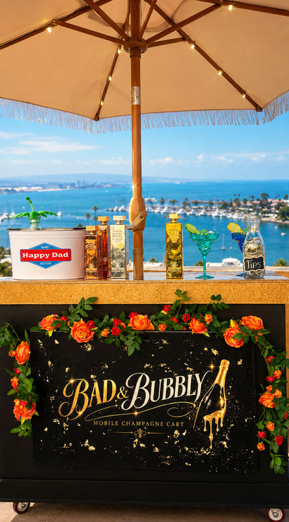 Rooftop mobile champagne cart decorated with orange roses and colorful bottles under a fringed umbrella, overlooking a sunny marina, harbor boats and a coastal lighthouse