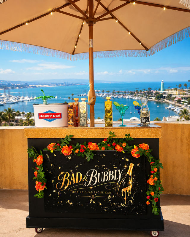 Rooftop mobile champagne cart decorated with orange roses and colorful bottles under a fringed umbrella, overlooking a sunny marina, harbor boats and a coastal lighthouse